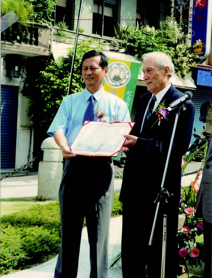 圖為 2001 年 6 月 2 日為紀念馬偕逝世百週年,偕約翰博士代表馬偕家族在馬偕博士雕像前舉行追思會,郭哲道鎮長頒給他淡水鎮永久榮譽鎮民,並當場宣佈每年 6 月 2 日為淡水馬偕日。